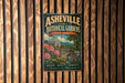 A vintage-style metal sign for the Asheville Botanical Gardens in North Carolina, featuring a scenic landscape with a greenhouse and flowers.