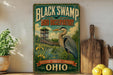 a vintage-style sign for the Black Swamp Bird Observatory, which is located in Ohio. The sign is displayed on a wooden shelf, surrounded by various kitchen utensils and a potted plant.