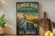 A vintage-style metal sign for the Bombay Hook National Wildlife Refuge in Delaware, featuring an illustration of a heron and a sunset over a body of water.