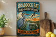 A vintage-style metal sign for the Braddock Bay Bird Observatory in New York, featuring an illustration of a blue heron and a sign that says "Discover, Protect, Educate" in a rustic style.