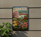 A vintage-style metal sign for "Brookside Gardens Maryland" is displayed on a wall, featuring a garden scene with colorful flowers and a building.
