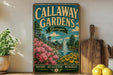 A vintage metal sign for Callaway Gardens in Georgia, featuring a scenic landscape with a fountain and flowers, is displayed on a shelf next to a wooden cutting board and some potted plants.