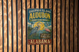 A vintage metal sign for Dauphin Island Audubon Bird Sanctuary in Alabama, featuring a blue heron and a bird in a grassy field.