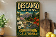 A vintage-style sign for Descanos Gardens, featuring a scenic landscape with a bridge, flowers, and mountains, is displayed on a shelf next to a potted plant and a cutting board.