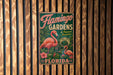 A vintage-style metal sign for Flamingo Gardens in Florida, featuring a pink flamingo and tropical foliage.