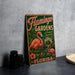 A vintage-style sign for Flamingo Gardens, featuring a pink flamingo and tropical foliage, is displayed on a gray wall.