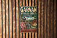 A vintage-style metal sign for the Garvan Woodland Gardens in Arkansas, featuring a scenic landscape with a bridge, flowers, and a waterfall.