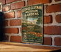A vintage-style metal sign for the George Eastman Museum Gardens in Rochester, New York, featuring an illustration of a garden with a fountain and a building in the background.