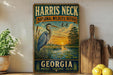 A vintage-style metal sign for the Harris Neck National Wildlife Refuge in Georgia, featuring an illustration of a heron and a sunset over a body of water.