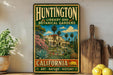 A vintage-style metal sign for Huntington Library and Botanical Gardens in California, featuring a tropical garden scene with palm trees, flowers, and a building.