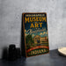 A vintage-style sign for the Indianapolis Museum of Art, featuring an illustration of the museum and its surroundings, is displayed on a gray concrete surface.