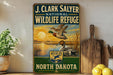 A vintage-style metal sign for the J. Clark Salyer National Wildlife Refuge in North Dakota, featuring a sunset landscape with ducks flying over a lake.