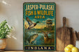 A vintage-style sign for the Jasper-Pulaski Fish & Wildlife Area in Indiana, featuring illustrations of a bird, a fish, and a deer in a natural setting.