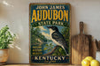 A vintage-style sign for John James Audubon State Park in Kentucky, featuring a black and white bird perched on a tree branch.