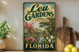 A vintage-style sign for Leu Gardens, a landmark in Orlando, Florida, featuring a tropical garden scene with palm trees, flowers, and a house.