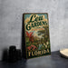 A vintage-style metal sign for Leu Gardens, located in Orlando, Florida, featuring a tropical garden scene with palm trees, flowers, and a house.