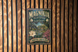 A vintage-style metal sign for Magnolia Plantation Gardens in South Carolina, featuring a lush garden scene with a large white flower and a house in the background.