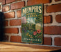A vintage-style metal sign for the Memphis Botanic Garden in Tennessee, featuring a lush garden scene with a greenhouse and various flowers.