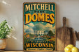 A vintage-style metal sign for Mitchell Park Domes in Milwaukee, Wisconsin, featuring a large illustration of the domed buildings and surrounded by a garden.