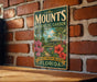 A vintage-style metal sign for the Mounts Botanical Garden in West Palm Beach, Florida, featuring a tropical garden scene with palm trees, flowers, and a building.