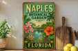 A vintage-style metal sign for the Naples Botanical Garden in Florida, featuring a tropical landscape with palm trees, flowers, and a body of water.