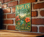 A vintage-style metal sign for the Naples Botanical Garden in Florida, featuring a tropical landscape with palm trees, flowers, and a pond.