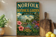 a vintage-style metal sign for the Norfolk Botanical Garden in Virginia, with a lush garden scene and a greenhouse visible. The sign is placed on a wooden shelf, surrounded by various kitchen utensils and a potted plant.