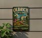A vintage-style metal sign for Olbrich Botanical Gardens in Madison, Wisconsin, featuring a garden scene with a building and flowers.