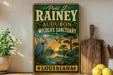 A vintage-style sign for Paul J. Rainey Audubon Wildlife Sanctuary in Louisiana, featuring an illustration of a heron and alligator in a pond with a sunset in the background.