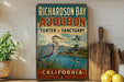 A vintage-style sign for "Richardson Bay Audubon Center & Sanctuary" in California, featuring an illustration of a heron and a lake.
