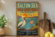 A vintage-style metal sign for the Salton Sea Bird Sanctuary in California, featuring illustrations of a pelican and flamingos against a sunset backdrop.