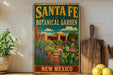 A vintage-style metal sign for Santa Fe Botanical Garden in New Mexico, featuring a colorful illustration of a desert landscape with cacti, flowers, and a building.