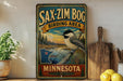 A vintage-style metal sign for the Sax-Zim Bog Birding Area in Minnesota, featuring a black and white bird perched on a pine tree branch.