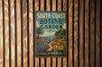 A vintage-style metal sign for the South Coast Botanic Garden in California, featuring a tropical garden scene with palm trees, flowers, and a winding path.