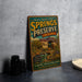 A vintage-style metal sign for Springs Preserve, Nevada, featuring a scenic landscape with mountains and a building, is displayed on a gray countertop next to a stack of black camera lenses.