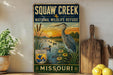 A vintage-style sign for Squaw Creek National Wildlife Refuge in Missouri, featuring an illustration of a heron and ducks in a pond with a sunset in the background.