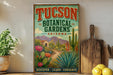 A vintage-style poster of the Tucson Botanical Gardens in Arizona, featuring a desert landscape with cacti, succulents, and colorful flowers. The poster is displayed on a wall, with a wooden cutting board and a potted plant to the left.