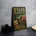 A vintage-style metal sign for " Tyler Arboretum" is displayed on a gray countertop, featuring a scenic landscape with a bridge and flowers.
