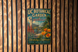 A vintage-style metal sign for UC Botanical Garden in Berkeley, California, featuring a tropical garden scene with palm trees, flowers, and a clock tower.