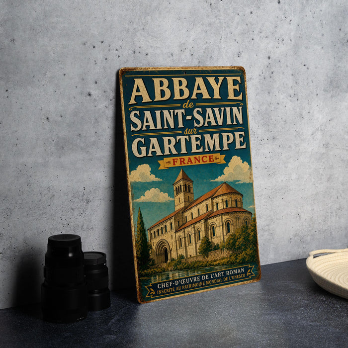 A vintage-style metal sign with the text "Abbaye de SAINT-SAVIN et GARTEMPE - FRANCE" and an illustration of a church, placed on a gray countertop next to a stack of black camera lenses.
