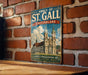 A vintage-style metal sign displaying the Abbey of St. Gall in Switzerland, with a brick wall in the background.