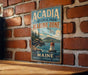 A vintage metal sign for Acadia National Park's Marine Zone, featuring a lighthouse and a seagull, is displayed on a brick wall.