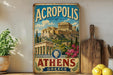 A vintage-style metal sign displaying the name "ACROPOLIS of Athens" and an illustration of the Acropolis, with a potted plant and wooden cutting board in the foreground.