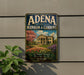 A vintage-style sign for "Adena mansion & gardens" is displayed on a wall, featuring a large illustration of a mansion surrounded by a well-maintained garden.