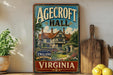 A vintage-style sign for Agedcroft Hall in Virginia, featuring a house surrounded by a garden, is displayed on a shelf next to a wooden cutting board and some lemons.
