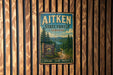 A vintage metal sign for Aitken State Forest in Vermont, featuring a scenic landscape with pine trees and a winding road.