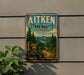 A vintage metal sign for Aitken State Forest in Vermont, featuring a scenic landscape with pine trees, a winding dirt road, and mountains in the background.