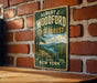 A vintage-style metal sign for Albert J. Woodford State Forest, featuring a scenic landscape with pine trees, mountains, and a lake.