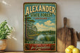 A vintage-style sign for Alexander State Forest in Louisiana, featuring a scenic image of a forest and a lake, is displayed on a shelf next to a wooden cutting board and some lemons.