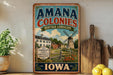 a vintage-style sign for "Amana Colonies Heritage Landscape" in Iowa, with a windmill and other rural scenery. The sign is displayed on a shelf next to a wooden cutting board and a potted plant.